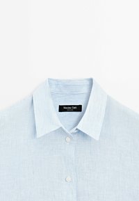 Camicia a righe azzurro chiaro con una texture morbida, chiusura a bottoni e colletto appuntito. L'etichetta riporta "Massimo Dutti" con dettagli di produzione visibili.