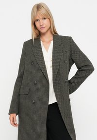 Dvojradový houndstooth blazer v tmavošedej a čiernej farbe so širokými klopami, gombíkovými detailmi a strihom na mieru. Nosený cez biele tričko.
