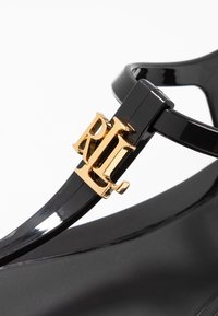 Svart glansig sandal med en gyllene logotypdetalj, med en stilren, minimalistisk design och ett tunt strap som kopplar tån till sulan.