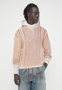 Pull en maille crème avec des rayures rouges, doté d'une encolure zippée et d'une capuche grise en dessous. Texture en maille lâche avec des poignets côtelés.