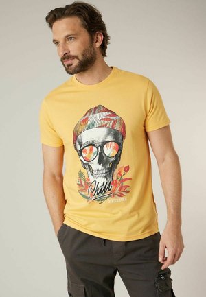 Man in een geel T-shirt met een kleurrijke schedelafbeelding die een zonnebril en een muts draagt, gecombineerd met donkere cargobroeken, staand met zijn hand in zijn zak.