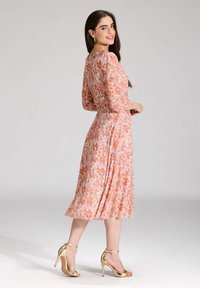 Kukallinen midi-mekko pitkillä hihoilla, vaaleanpunaisen ja oranssin sävyissä, jossa on muotoiltu yläosa ja laskostettu hame. Yhdistetty kultaisiin remmikorkokenkiin.