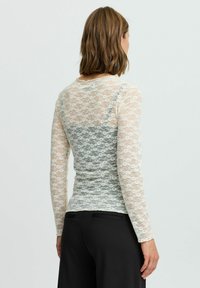 Top en dentelle à manches longues avec un motif floral, couleur crème clair, et design ajusté, superposé sur une couche unie bleu clair.