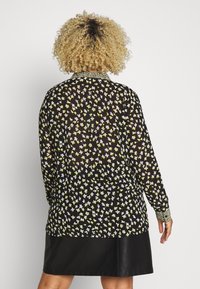 Blouse noire transparente avec imprimé floral jaune et blanc, manches longues et col orné de décorations. Portée avec une jupe en cuir noire.