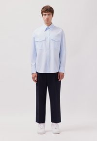 Camicetta azzurro chiaro a maniche lunghe con due tasche frontali, polsini con bottoni, abbinata a pantaloni navy e sneakers bianche. Design semplice e strutturato.