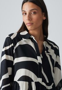 Schwarz-weiße gestreifte Bluse mit Kragen, versehen mit einer Knopfleiste und gerafftem Stoff an der Taille. Leichter, glatter Stoff.