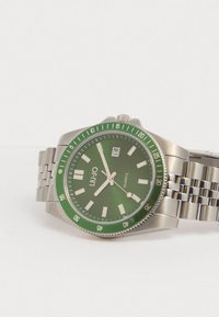 Kvartsikello, jossa on vihreä kellotaulu valkoisilla tuntimerkeillä, hopeametallinen kotelo ja ranneke sekä vihreä bezel minuuttimerkeillä.