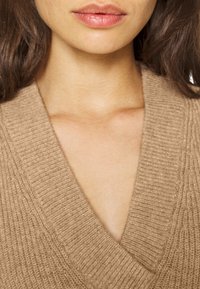 Beige gebreide trui met een diep V-hals en een gestructureerd ribpatroon, met een zachte, warme stof. Geen extra patronen of metaaldetails aanwezig.
