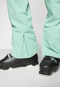 Pantalones de esquí impermeables en verde menta con aberturas con cremallera en los tobillos, combinados con botas de esquí negras con acentos blancos y hardware ajustable.