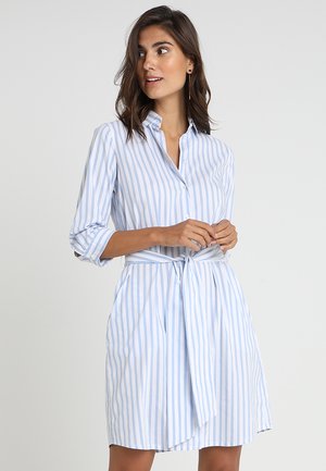 Donna che indossa un vestito camicia a righe verticali azzurre e bianche con maniche 3/4 e cintura annodata in vita, che guarda di lato.