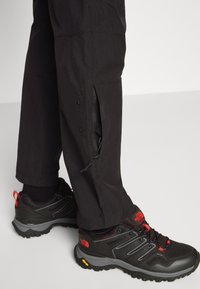 Svarta utomhusbyxor med dragkedjade sidoventiler, i kombination med svarta vandringsskor med röda accenter och strukturerade sulor.