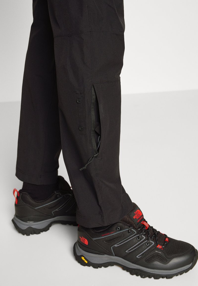 Svarta utomhusbyxor med dragkedjade sidoventiler, i kombination med svarta vandringsskor med röda accenter och strukturerade sulor.