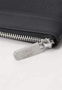 Črn usnjen etui z zlatim zadrgo in oznako. Ima gladek zgornji del in pleten spodnji del. Oblikoval Karl Lagerfeld za Disney.