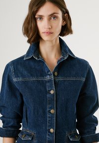 Denim shirt in dark blue with a collared neckline, button front, and chest pocket. Features rolled sleeves and contrasting stitching details.