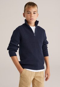 Marinblått ribbat stickat tröja med halvmatchande krage, parat med en vit t-shirt. Texturerad tyg, uppvikta ärmar och avslappnad passform.