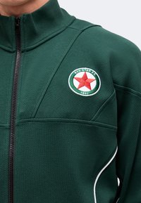 Πράσινο αθλητικό μπουφάν με μαύρο φερμουάρ και στρογγυλό έμβλημα Red Star FC με κόκκινο αστέρι και το έτος 1897 στο αριστερό στήθος.