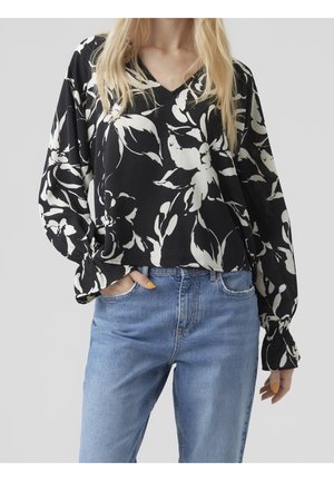Woman wearing black long-sleeve blouse with large white floral print and light blue high-waisted jeans, hand in pocket.