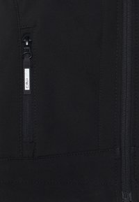 Black softshell jacket featuring a zippered pocket with branded pull tab, textured fabric, and visible stitching details.