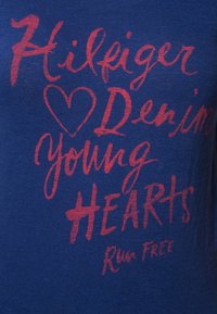 Blå bomullst-shirt med röd grafisk text som lyder "Hilfiger Denim Young Hearts Run Free" i olika typsnitt och storlekar.