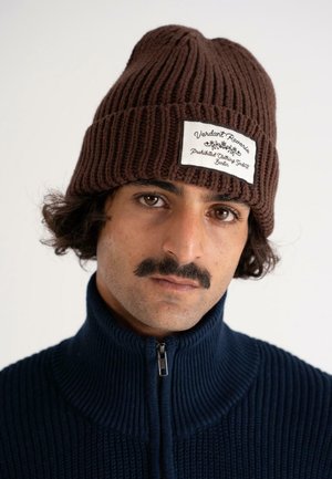 Un homme avec une moustache porte un bonnet en tricot marron avec une étiquette blanche et un sweat zippé bleu marine, le tout sur un fond clair et uni.