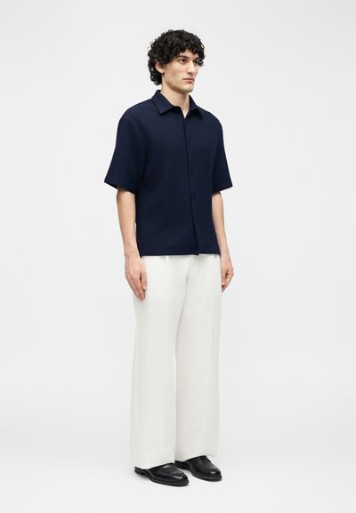 Άνδρας μοντέλο στέκεται φορώντας μπλε πουκάμισο με κοντά μανίκια και κουμπιά, άσπρο παντελόνι με φαρδιά μπατζάκια και μαύρα δερμάτινα loafers, πάνω σε απλό φόντο.