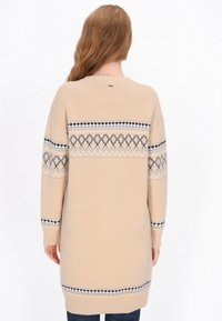 Beige neulepusero, jossa on rento istuvuus ja yläselän sekä hihojen kohdalla kuvioitu sinivalkomusta nauha.