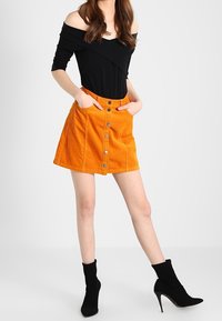 Haut noir à épaules dénudées avec une jupe trapèze en velours côtelé orange vif, dotée d'une fermeture boutonnée à l'avant et de poches latérales, assortie à des bottines noires.