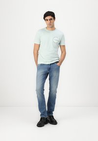 T-shirt di cotone verde chiaro con scollatura rotonda e dettaglio del logo stampato sul lato sinistro, abbinata a jeans in denim blu e scarpe nere.