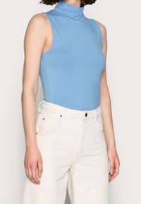 Light blue sleeveless turtleneck bodysuit in smooth fabric, paired with high-waisted white jeans featuring front pockets and a button closure.