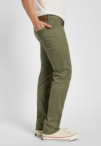 Jeans verde oliva con corte slim, detalles de costura sutil y una etiqueta de cinturón de cuero marrón. Combinados con zapatillas beige.