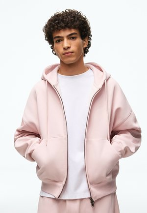 Jeune homme aux cheveux bouclés portant un sweat à capuche zippé rose clair sur un t-shirt blanc, les mains dans les poches, debout devant un fond uni.
