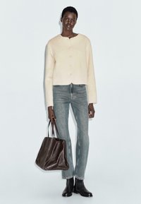 Donna che indossa un cardigan crema con bottoni, jeans grigi, stivali neri, mentre tiene una grande borsa a spalla in pelle marrone scuro, in piedi contro uno sfondo bianco semplice.