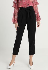 Pantalones negros ajustados con cintura ceñida, de longitud hasta el tobillo y textura suave, combinados con una blusa de manga larga floral y tacones blancos.