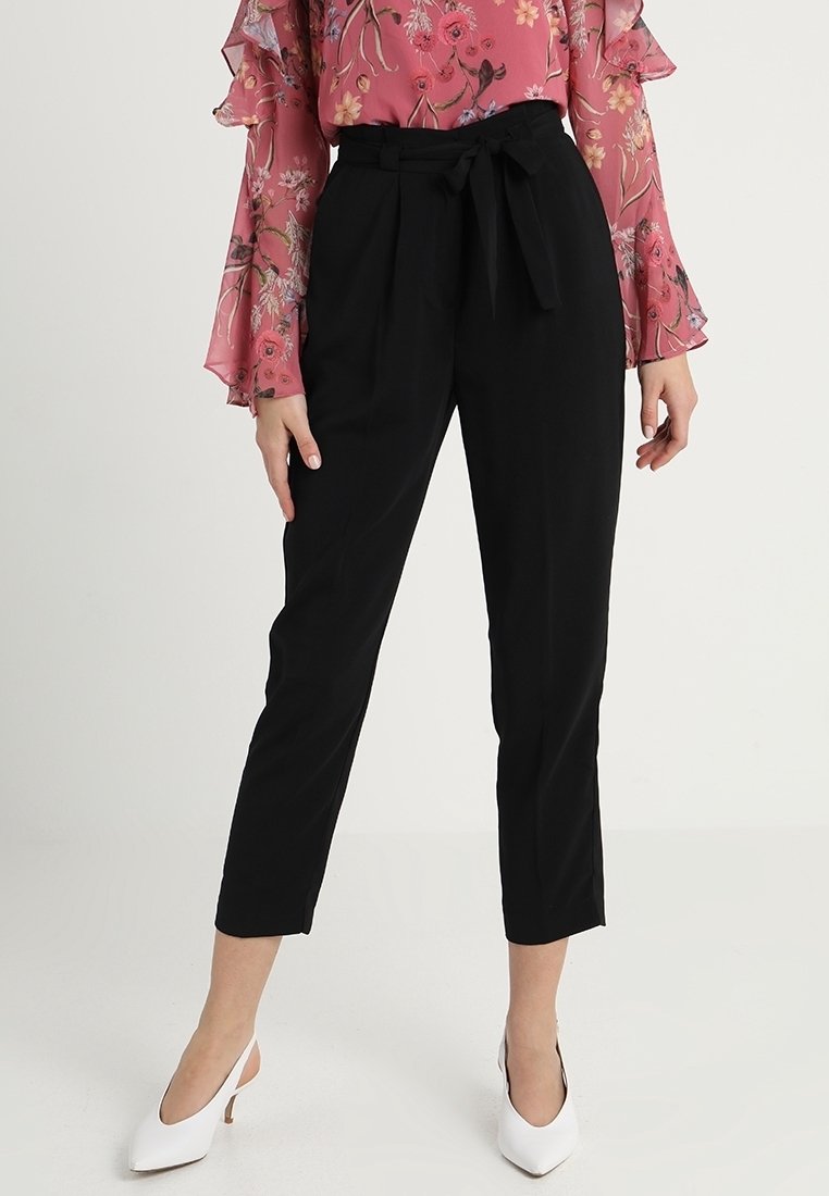 Pantalones negros ajustados con cintura ceñida, de longitud hasta el tobillo y textura suave, combinados con una blusa de manga larga floral y tacones blancos.
