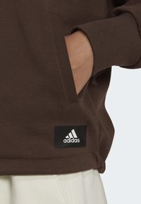 Sudadera marrón con puños acanalados y un bolsillo lateral. Presenta una etiqueta del logo negro de adidas cerca del dobladillo. Tela suave y texturizada.