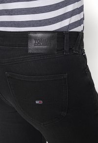 Vaqueros de mezclilla negros con una etiqueta de cuero de TOMMY JEANS en la parte trasera de la cintura, con un pequeño logo bordado en el bolsillo derecho. Camisa de rayas visible arriba.