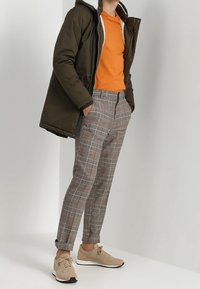 Parka verde oliva con forro blanco, camiseta naranja, pantalones de cuadros grises y zapatillas beige con diseño de cordones y sutiles acentos.