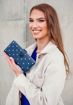 Navy blue wallet with white polka dots, made of textured material. Fastened with a button, featuring a circular logo on the front.