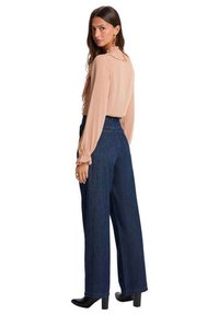 Mujer con blusa rosa claro con volantes, vaqueros de pierna ancha azul oscuro de talle alto y botas negras de tacón, de pie y mirando por encima del hombro.