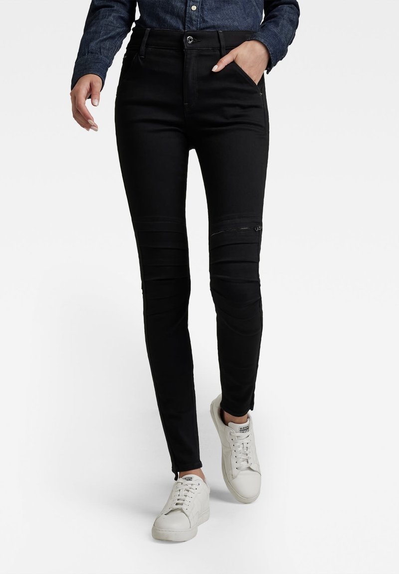 Personne portant un pantalon noir ajusté avec des nervures au niveau des genoux et un détail zippé, associé à des baskets blanches et une chemise à manches longues bleu foncé.
