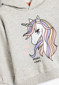 Felpa grigia con cappuccio caratterizzata da una grafica di unicorno. L'unicorno ha una criniera rosa, viola e gialla con cuori e il testo "Vibrazioni magiche".