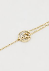 Kuldne käevõru ümmarguse ripatsi ja selge rhinestone kaunistustega, millel on brändi nimi "GUESS" kalligraafilises kirjas.