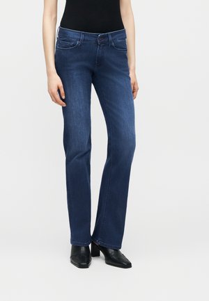 Személy középig érő, sötétkék, egyenes szárú farmerben, fekete felsőben és fekete magassarkú cipőben, áll egy egyszerű, világos háttér előtt.