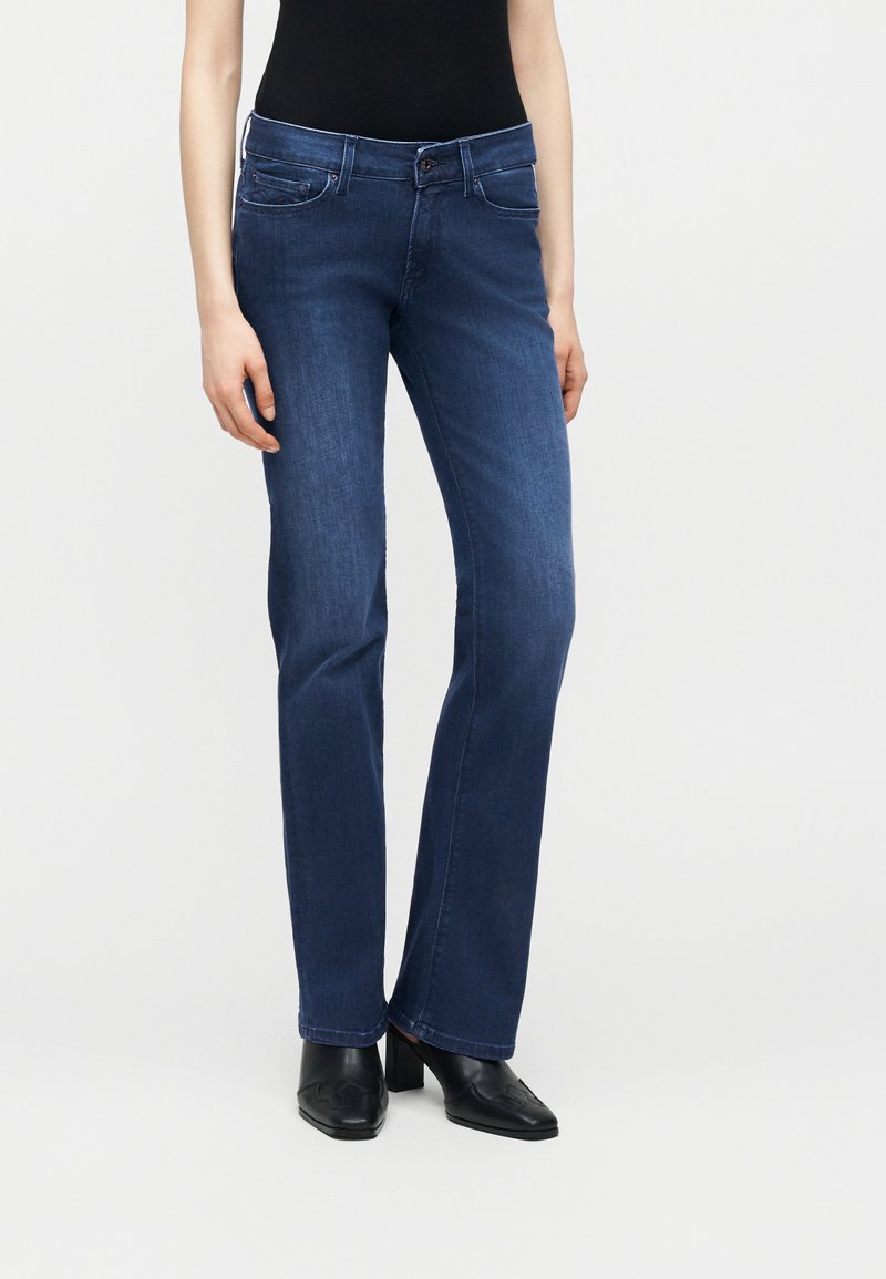 Άτομο που φορά μεσαίου ύψους σκουρό μπλε τζιν στενή γραμμή, μαύρο τοπ και μαύρα παπούτσια με τακούνι, στέκεται μπροστά σε απλό φωτεινό φόντο.