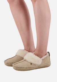 Beige pantoffels van suède, gevoerd met crèmekleurige imitatiebont. Zachte pluche manchetten en decoratieve stiksels langs de randen. Platte zolen.