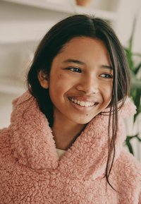 Rosa, flauschiger Fleece-Hoodie mit großem Kragen. Weiche Textur, lockere Passform. Einfaches weißes Oberteil darunter, innere Umgebung mit Grünpflanzen.