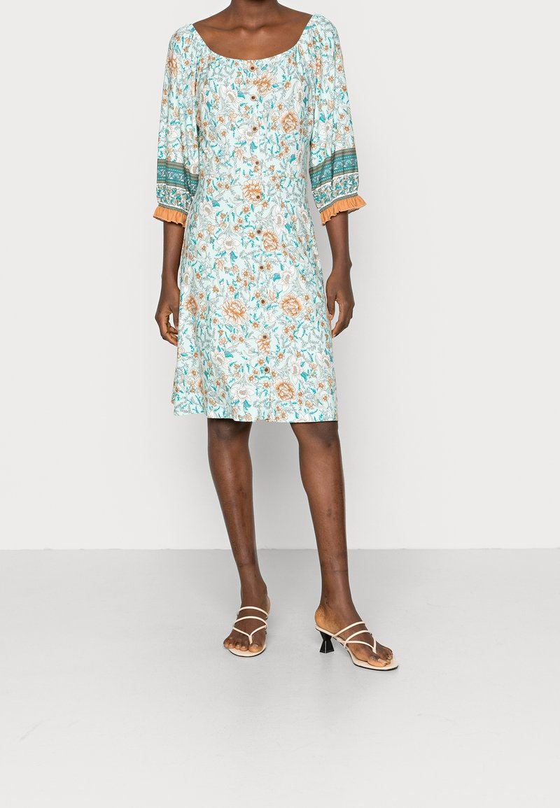 Blå blommig klänning med fyrkantig halsringning och puffärmar, med orange detaljer. Kombineras med beiga sandaler med remmar.