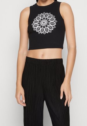 Woman wearing a black sleeveless crop top with a white floral mandala design, paired with high-waisted black pleated pants.