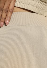 Beige žebrovaná tkanina s hladkým povrchem, ukazující pás a ruku odpočívající na něm. Materiál se zdá být elastický a přiléhavý.