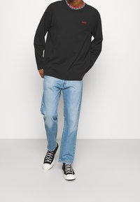 Schwarzes Langarmhemd mit roten bestickten Text, kombiniert mit hellblauen Jeans und schwarzen High-Top-Sneakern.
